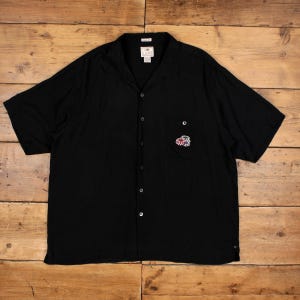 May include: Black short-sleeve button-down shirt with a small embroidered design on the chest pocket. The shirt has a classic collar and a relaxed fit. The shirt is made of a lightweight fabric.