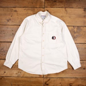 May include: Off-white button-down shirt with long sleeves. Features a small pocket on the left chest with a maroon and black logo. The shirt has a button-down collar and is made of a soft, likely cotton, material. A classic, casual shirt.