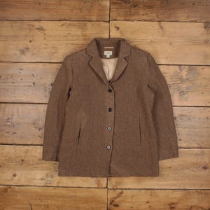 Puede incluir: Un blazer marrón con un patrón texturizado. El blazer tiene solapas con muescas, tres botones y dos bolsillos delanteros. El forro es de color beige claro. El blazer se muestra sobre una superficie de madera.