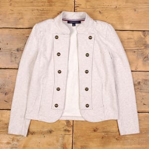 May include: A light grey jacket with a stand-up collar and double-breasted button closure. The jacket features eight gold-coloured buttons and a white interior lining. The Tommy Hilfiger logo is visible on the collar.