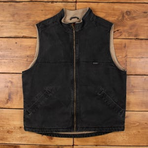 May include: A black vest with a full-length zipper and a beige, fuzzy interior lining. The vest has two front pockets and a stand-up collar. The exterior is a dark, faded black color. The vest is made of a durable material.