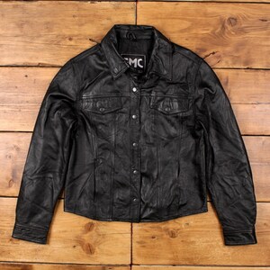 May include: Black leather jacket with a button-up front, two chest pockets, and a collared neckline. The jacket has a classic design and is made of a smooth, textured leather. The jacket is displayed on a wooden surface.