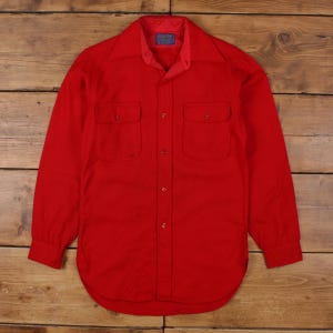 Puede incluir: Una camisa roja brillante de manga larga con dos bolsillos en el pecho y cierre de botones. La camisa tiene un cuello clásico y un dobladillo redondeado. La tela parece gruesa, posiblemente de lana. La camisa se muestra sobre una superficie de madera.