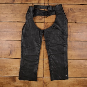 Può includere: Chaps da moto in pelle nera con cintura e fibbia. I chaps hanno una tasca sulla gamba destra e chiusure a scatto sui lati. La pelle appare usurata, suggerendo uno stile vintage.