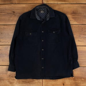 May include: A navy blue long-sleeved shirt with two chest pockets and a button-down front. The collar has a striped pattern. The shirt is made of a thick, durable material, suitable for cooler weather.