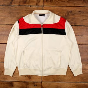 May include: Cream-colored track jacket with a full zipper, featuring a red and black stripe design across the chest. The jacket has a stand-up collar, ribbed cuffs, and a ribbed hem. The jacket has two front pockets.