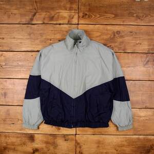 Puede incluir: Chaqueta gris y azul marino con cremallera y diseño de chevrón. La chaqueta tiene cuello alto, puños elásticos y dobladillo con cordón. La chaqueta está hecha de un material ligero.