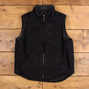 Puede incluir: Un chaleco negro sin mangas con cremallera frontal completa, cuello alto y forro de forro polar estilo sherpa en gris oscuro. La prenda incluye un bolsillo vertical con cremallera en el pecho izquierdo y dos bolsillos abiertos en la cintura.