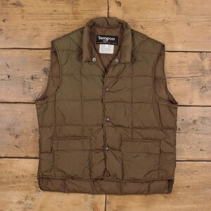 May include: Olive green quilted vest with a button-up front and two front pockets. The vest has a collar and the brand name "Tempco" is visible on the label. The vest is made of a quilted material.