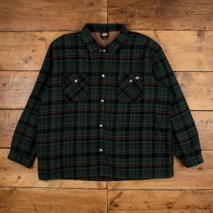 May include: A long-sleeve button-up shirt featuring a dark green and black plaid pattern with thin brown accent lines. The shirt includes a pointed collar, two chest pockets with snap closures, and a brown interior lining with a Dickies brand label.