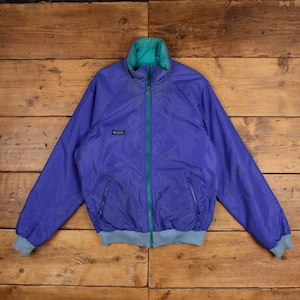 Può includere: Una giacca Columbia viola con colletto alto, caratterizzata da una fodera interna color ottanio e una cerniera frontale completa dello stesso colore. La giacca presenta due tasche laterali con zip, maniche lunghe con polsini a coste azzurri.