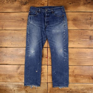 May include: A pair of blue denim jeans featuring a button fly closure, five pockets, and a distressed finish with light fading on the thighs and knees. The hem is frayed, and there are small white paint-like splatters across the legs.