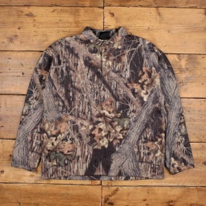 Pode incluir: Uma camisa de lã polar camuflada de manga comprida com gola abotoada. A camisa apresenta um padrão de galhos e folhas de árvores marrons, verdes e bege. A peça de roupa está colocada sobre uma superfície de madeira.
