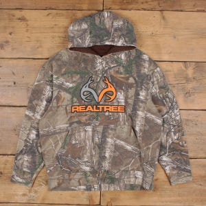Könnte beinhalten: Ein Kapuzenpullover mit Camouflage-Muster, Kapuze und Vordertasche. Der Kapuzenpullover zeigt das "Realtree"-Logo in Orange und Grau. Der Hintergrund ist eine Holzoberfläche.