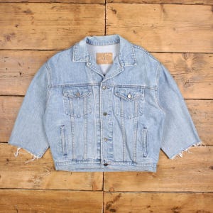 May include: Light blue denim jacket with short sleeves. The jacket has a classic design with two chest pockets and two side pockets. It features a button-up front and a collar. The sleeves are cut off, giving it a unique, distressed look.