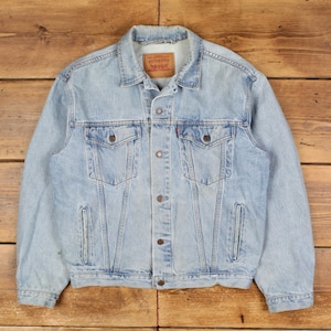 Könnte beinhalten: Hellblaue Jeansjacke mit zwei Brusttaschen und zwei Seitentaschen. Die Jacke hat eine Knopfleiste vorne und einen Kragen. Ein rotes und braunes Etikett ist auf der Rückseite des Kragens sichtbar. Die Jacke ist aus hellblauem Denim.