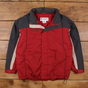Puede incluir: Chaqueta Columbia roja y gris con cuello alto y cierre de cremallera. La chaqueta tiene mangas y detalles grises, con una franja beige en los laterales. El logotipo de Columbia es visible en el pecho.