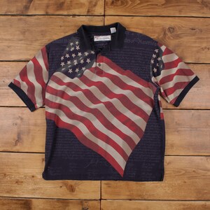 May include: A navy blue polo shirt with a large American flag design in red, white, and tan. The flag design covers the front and part of the sleeves. The shirt has a black collar and sleeves. The text "American Spirit" is visible.