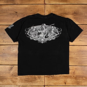 Peut inclure: T-shirt noir avec un motif graphique au dos représentant trois femmes en uniforme militaire, le texte "AMERICAN SPECIAL OPS" et un crâne. La manche gauche présente un motif à rayures blanches.