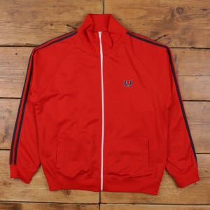May include: A red track jacket with a white zipper and navy blue stripes down the sleeves. The jacket has a small emblem on the chest and two pockets. The jacket is made of a smooth, shiny material.