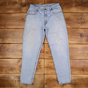 May include: Light blue denim jeans with a classic five-pocket design. The jeans have a straight leg cut and a button and zipper closure. The denim has a faded appearance, with some wear and tear visible. The jeans are laid flat on a wooden surface.