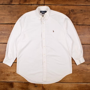 Può includere: Camicia bianca a maniche lunghe con colletto button-down. La camicia presenta un piccolo logo marrone sul petto. La camicia è realizzata in un tessuto morbido e tessuto.