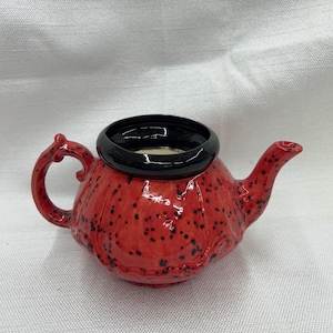 May include: A small, red ceramic teapot with a black rim and handle. The teapot has a textured surface with black speckles. The spout and handle are curved. The teapot is sitting on a white surface.