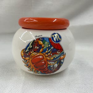 May include: A white ceramic jar with an orange lid. The jar features a colorful illustration of the Scorpio zodiac sign, including a scorpion and the word "Scorpio". The lid is a solid orange color.