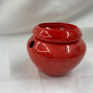 May include: A small, red ceramic container with a lid. The container has a rounded body and a circular opening at the top. The red glaze has small black speckles. There is a small hole on the side of the container.