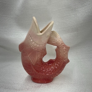 May include: A ceramic fish-shaped vase in shades of pink and red. The vase has an open mouth and a curved tail, with a textured scale pattern. The vase is a decorative item.