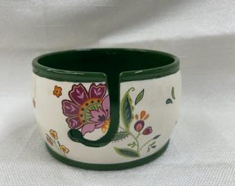 May include: A white ceramic napkin holder with a green rim and interior. The holder features a floral design with purple, pink, and orange flowers and green leaves. The holder is round with a curved opening.
