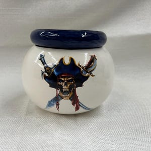 May include: A white ceramic jar with a dark blue lid. The jar features a pirate skull design with crossed swords, a tricorn hat, and red accents. The jar is round and appears to be a decorative item.