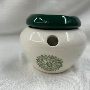 May include: A white ceramic oil warmer with a dark green lid. The warmer has a circular hole and a green floral design. The oil warmer is designed to hold essential oils and diffuse their fragrance.