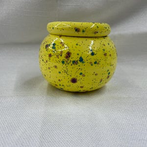 May include: A small, round, yellow ceramic jar with a matching lid. The jar is speckled with small dots of blue, green, and brown. The jar is sitting on a white surface.