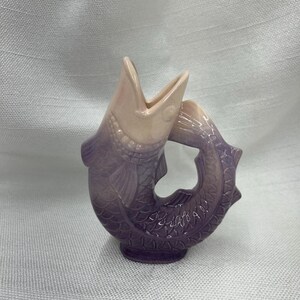 May include: A ceramic fish-shaped vase in shades of purple and pink. The vase has an open mouth and a tail that forms a handle. The fish has detailed scales and fins.