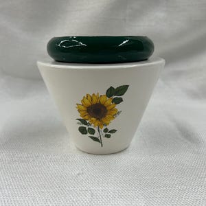 May include: A white ceramic container with a sunflower design, topped with a dark green, rounded lid. The sunflower is yellow and brown with green leaves. The container has a flared shape.
