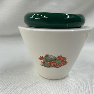 May include: A white ceramic container with a green lid. The container has a cactus and flower design. The lid is a solid, glossy green. The container is likely a decorative item or a small storage vessel.
