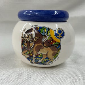 May include: A ceramic jar with a blue lid and a white body. The jar features a colorful illustration of a bull, the Taurus zodiac sign, with the text "Taurus" and the date "21.04.-20.05."
