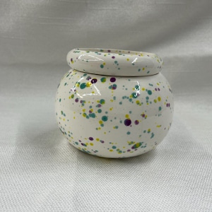 May include: A small, round, white ceramic jar with a matching lid. The jar and lid are decorated with a speckled pattern of teal, purple, and yellow dots. The jar is sitting on a white surface.