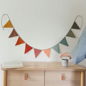 May include: A decorative bunting banner featuring a series of triangular fabric flags in a gradient of colors including mustard yellow, brown, orange, red, pink, peach, light blue, and sage green, suspended from a thin cord against a wall.