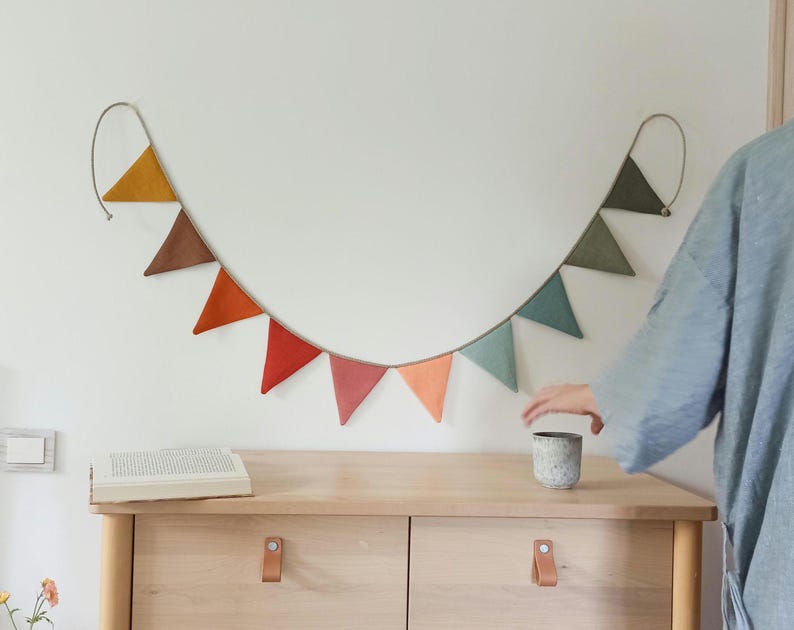 May include: A decorative bunting banner featuring a series of triangular fabric flags in a gradient of colors including mustard yellow, brown, orange, red, pink, peach, light blue, and sage green, suspended from a thin cord against a wall.
