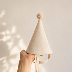 Può includere: Un cappello da festa conico di colore beige chiaro a tinta unita, caratterizzato da un piccolo pon-pon abbinato sulla punta. Il cappello include due lunghi nastri di tessuto piatto fissati alla base per fissare l'oggetto, realizzato in tessuto semplice.