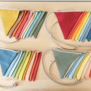 May include: Four sets of triangular fabric bunting flags arranged in groups. Each set features a variety of solid colors including yellow, red, blue, green, and orange, all attached to a thick, braided beige rope for hanging and display.