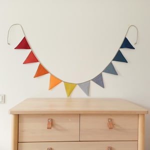 May include: A decorative bunting banner featuring a series of triangular fabric flags in a gradient of colors, transitioning from deep red and orange on the left to yellow, gray, and blue on the right, all suspended from a single light-colored cord.