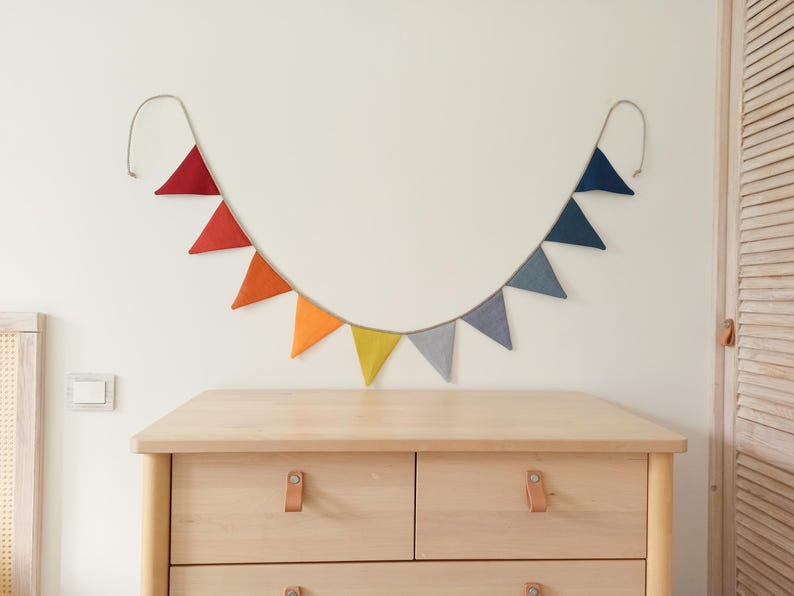 May include: A decorative bunting banner featuring a series of triangular fabric flags in a gradient of colors, transitioning from deep red and orange on the left to yellow, gray, and blue on the right, all suspended from a single light-colored cord.