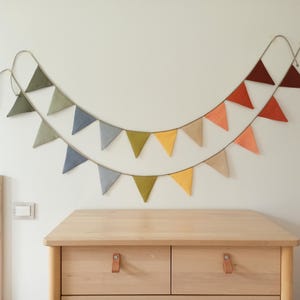 May include: A two-tiered fabric bunting banner featuring triangular flags in a gradient of colors including olive green, slate blue, mustard yellow, beige, peach, and deep rust, all suspended from a thin cord arranged in a curved shape.