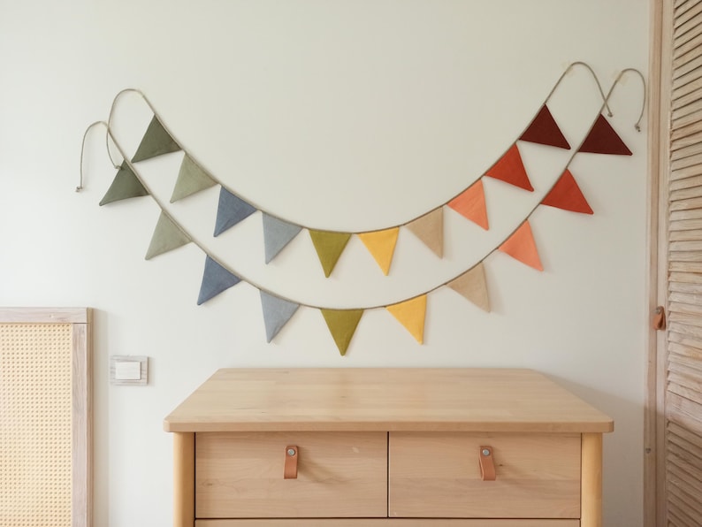 May include: A two-tiered fabric bunting banner featuring triangular flags in a gradient of colors including olive green, slate blue, mustard yellow, beige, peach, and deep rust, all suspended from a thin cord arranged in a curved shape.