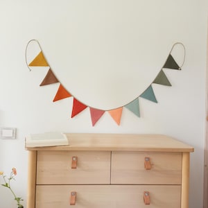 May include: A decorative bunting banner featuring ten triangular fabric flags in a gradient of colors including mustard yellow, brown, orange, red, pink, peach, light blue, sage green, and dark green, all suspended from a single thin cord.