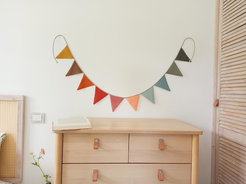 May include: A decorative bunting banner featuring ten triangular fabric flags in a gradient of colors including mustard yellow, brown, orange, red, pink, peach, light blue, sage green, and dark green, all suspended from a single thin cord.