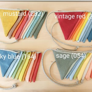 May include: Four sets of triangular fabric bunting flags attached to a braided rope. Each set features a gradient of colors, labeled as mustard (232), vintage red (338), sky blue (164), and sage (054), showcasing various textile hues.